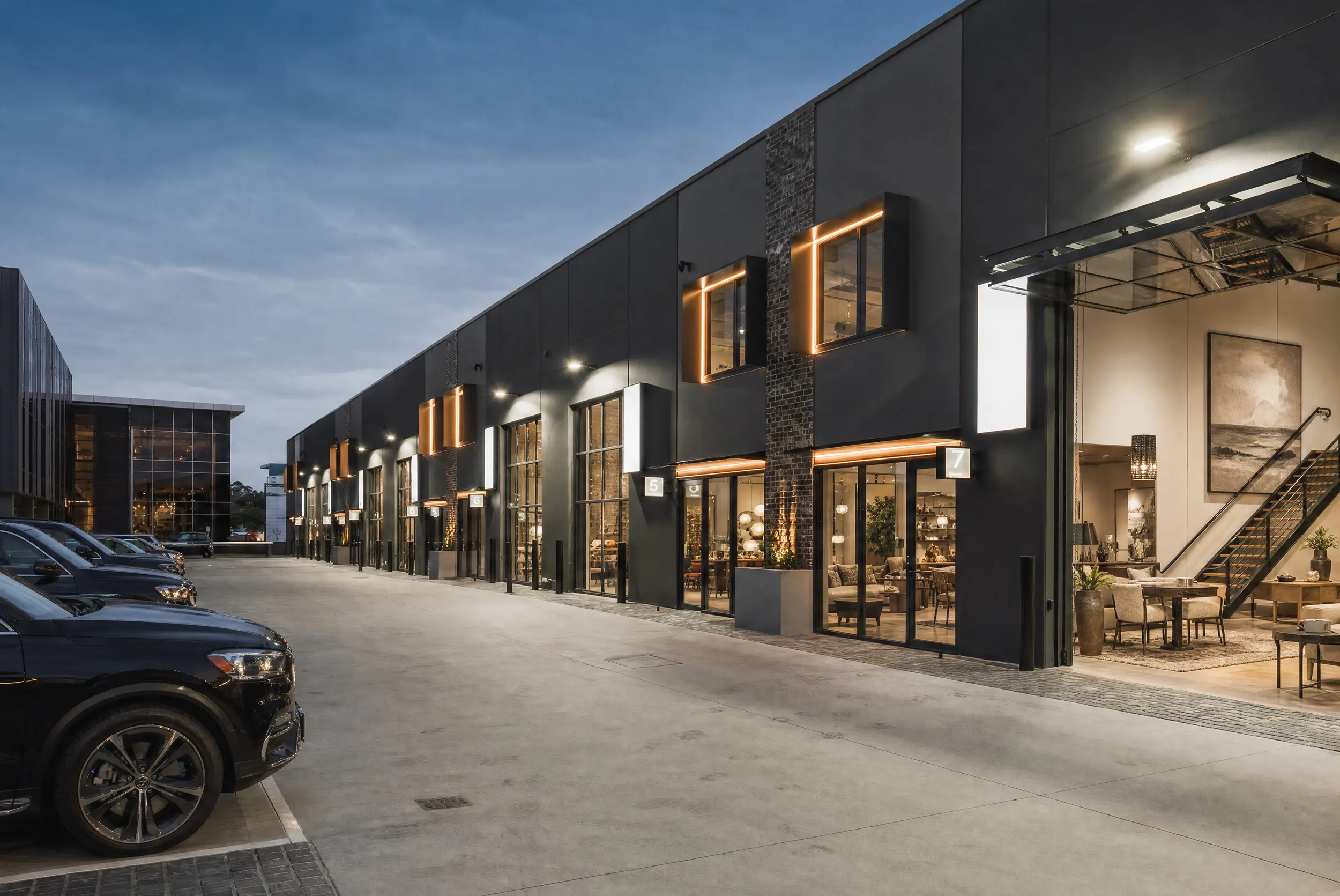 The Jacoah Place frontage at dusk — a row of numbered showroom tenancies with warm-lit glazing, full-height glass doors and dedicated parking