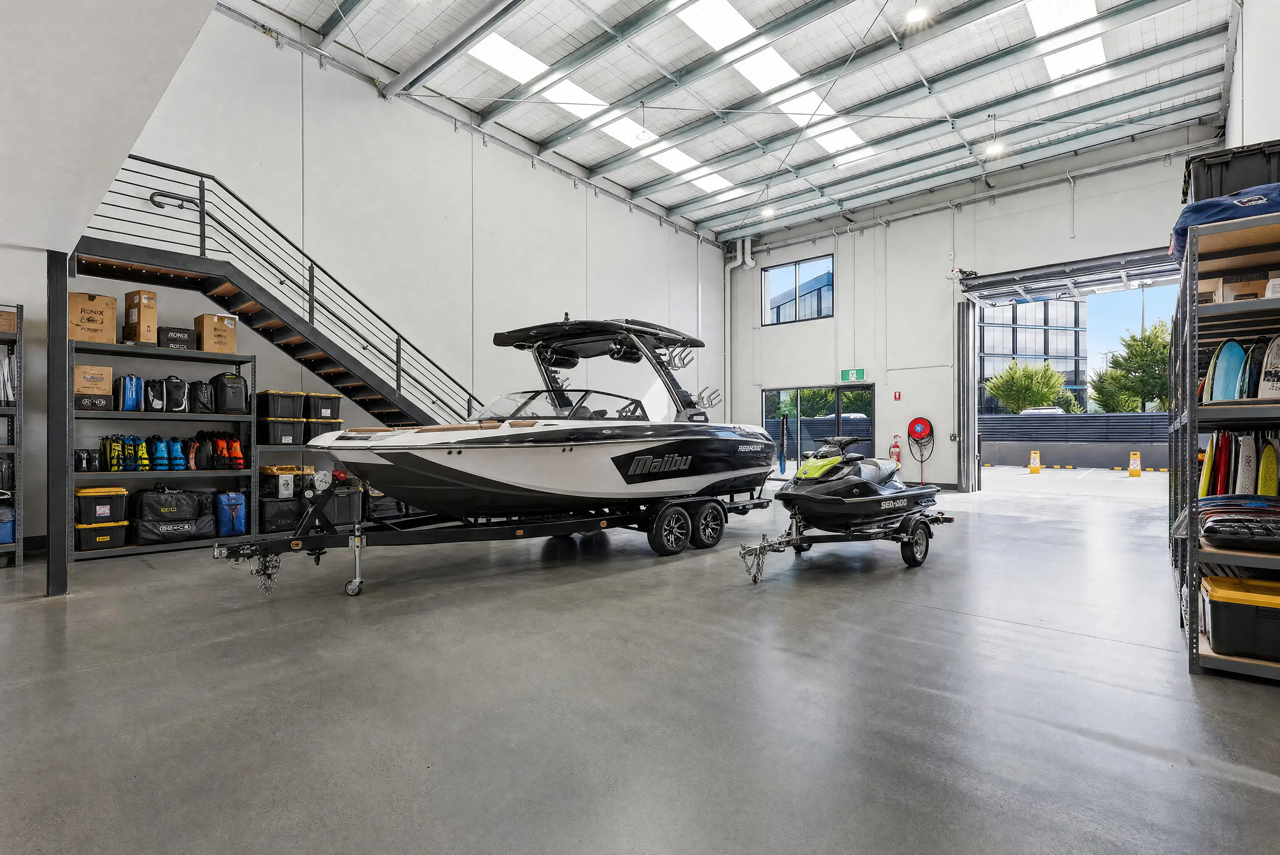A Jacoah Place tenancy styled for boat and jet-ski storage, with a ski boat on its trailer beside racked wakeboards and the roller door open to the driveway